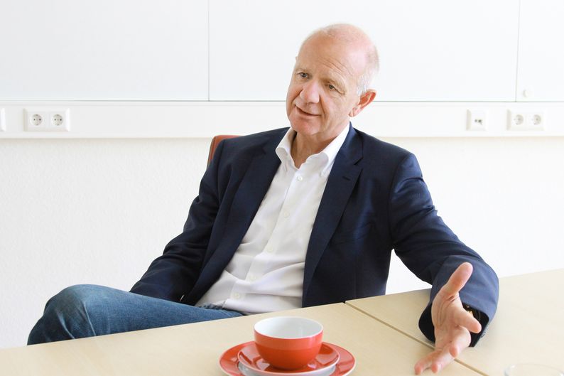 Ralf Fischer sitzt an einem Tisch und spricht mit offener Handbewegung.