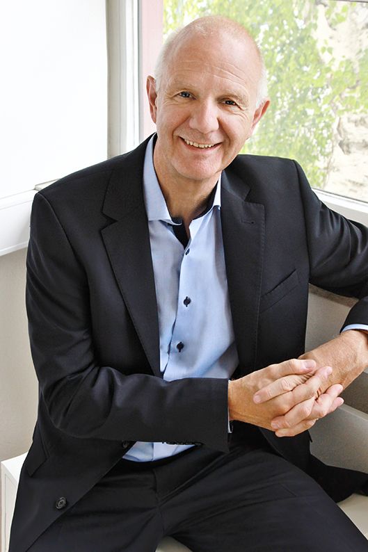 Ralf Fischer im dunklen Anzug, sitzend vor einem Fenster, die Hände locker gefaltet.